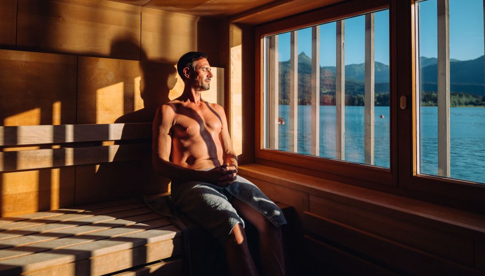 Mann sitzt in der Panorama-Sauna mit Blick auf den Wolfgangsee im Adults Only Hotel Cortisen am See im Salzkammergut
